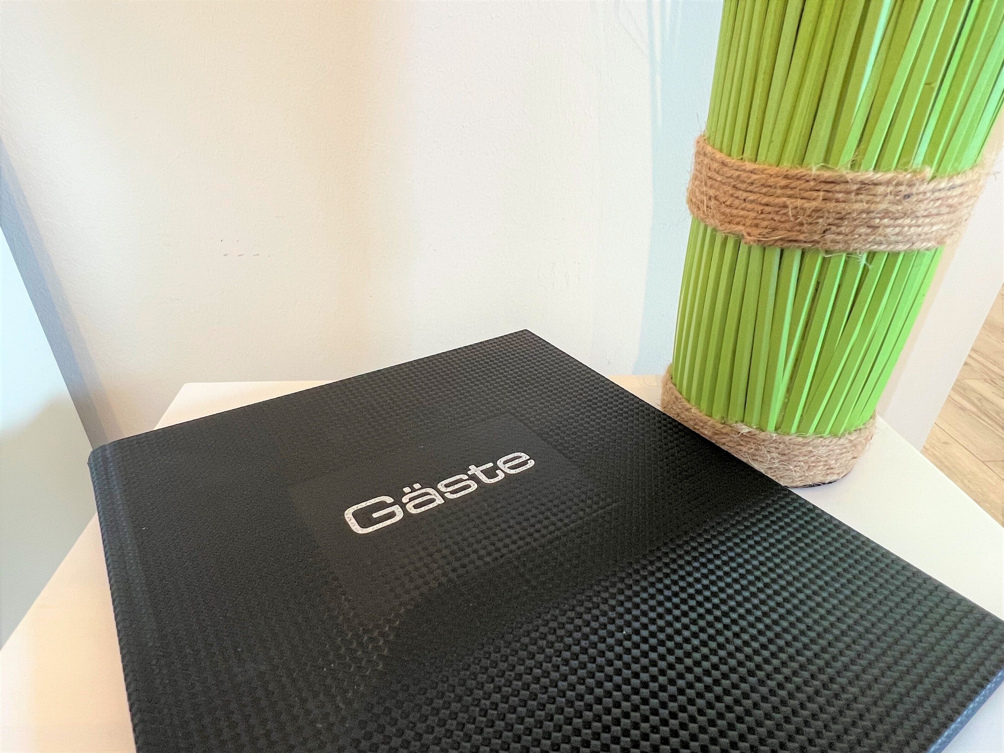 En sort kasse med ordet "Gäste" på, placeret på en hvid overflade ved siden af en grøn plante bundet med garn.