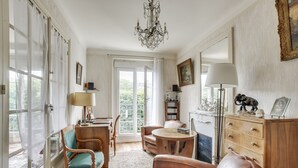 Uma sala de estar ampla, com um candelabro, lareira, um armário de madeira e uma grande janela com cortinas.