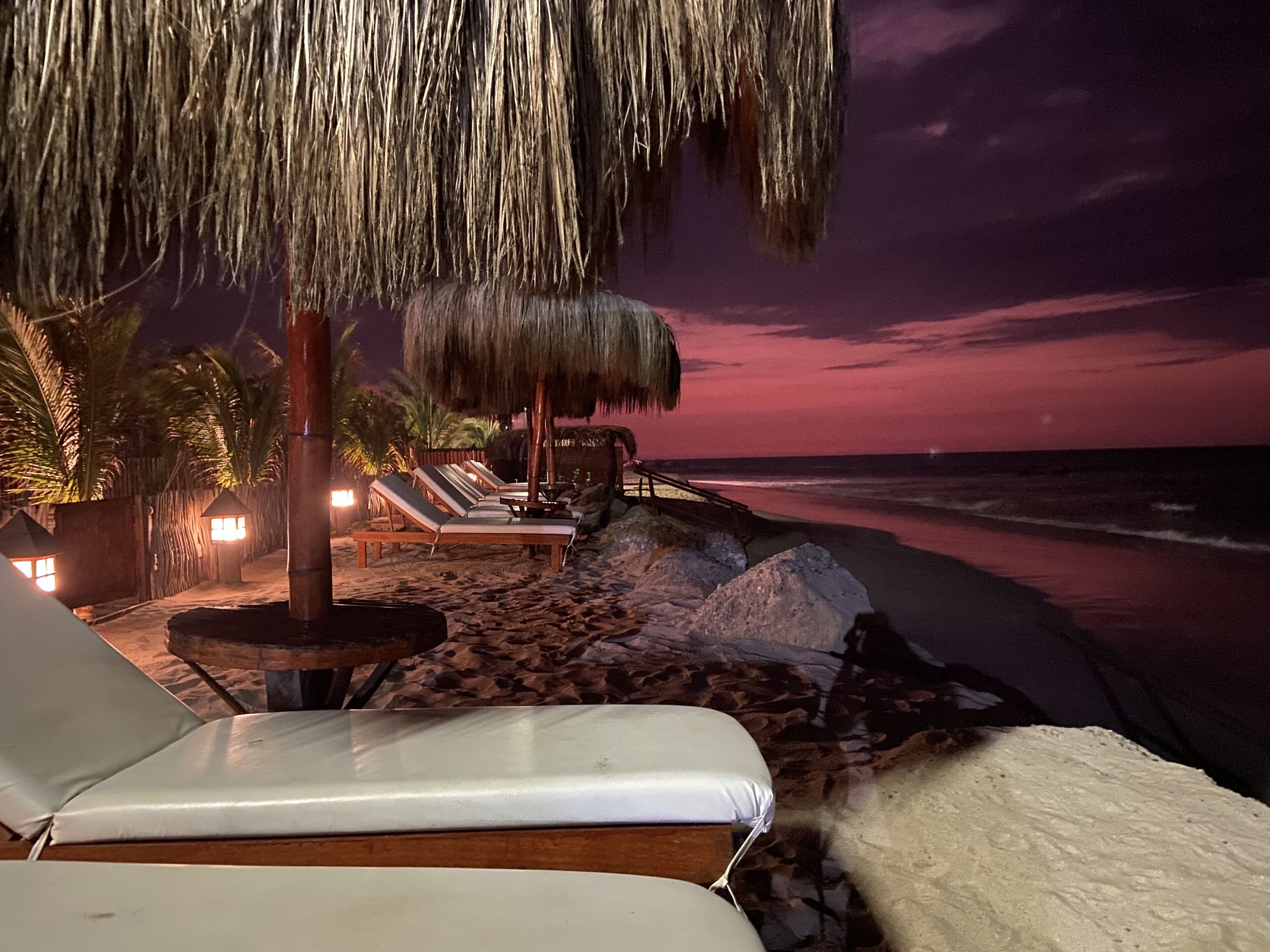 Un área de descanso en la playa con tumbonas y un paraguas de paja, con vista al océano al atardecer.