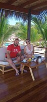 Una pareja sentada en un banco de madera con una mesa frente a ellos, que incluye una cafetera, tazas y un frutero.