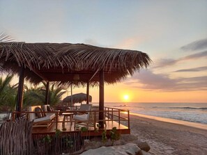 A beachside lounge with a thatched roof, wooden deck, and seating area overlooking the ocean at sunset. - El Samay Hotel Boutique (Canoas de Punta Sal)
