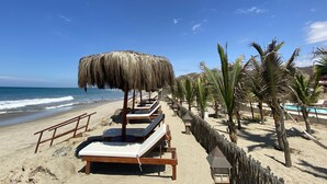 A beach with wooden lounge chairs and thatched umbrellas, a wooden fence, and palm trees. - El Samay Hotel Boutique (Canoas de Punta Sal)