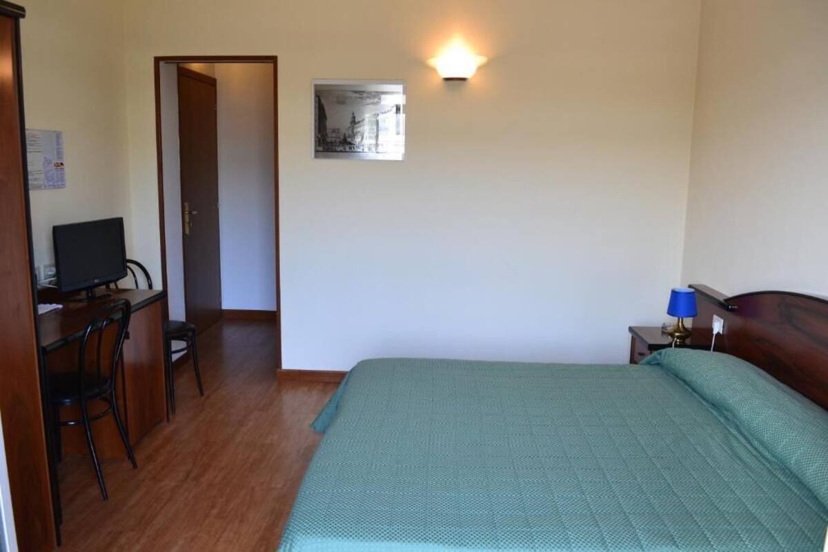 A bedroom with a bed, a desk with a chair, a wall-mounted light, and a picture on the wall.