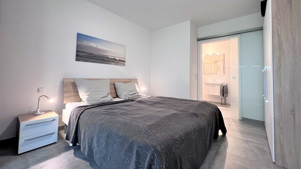 A modern bedroom with a large bed, a bedside table, a nightstand, a wall-mounted lamp, and a bathroom visible through an open door. - Hello and welcome to the "Lighthouse" (Dangast)