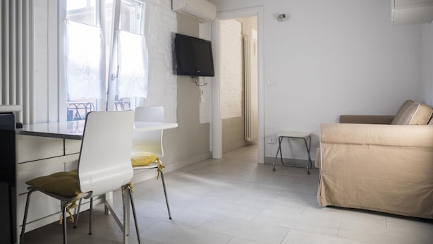 Een kamer met een wit bureau, een stoel, een beige bank, een aan de muur bevestigde televisie en een raam met zonwering.