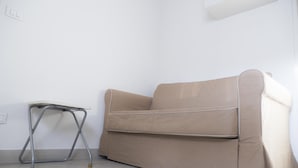 Een beige bank en een klein tafeltje met wielen in een kamer met witte muren en betegelde vloer.
