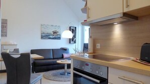 A modern kitchen with a built-in oven, a dining area with a black sofa and a small round table, and a painting of a house on the wall.