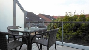 A balcony with a glass table and wicker chairs, overlooking a residential area with houses and greenery.