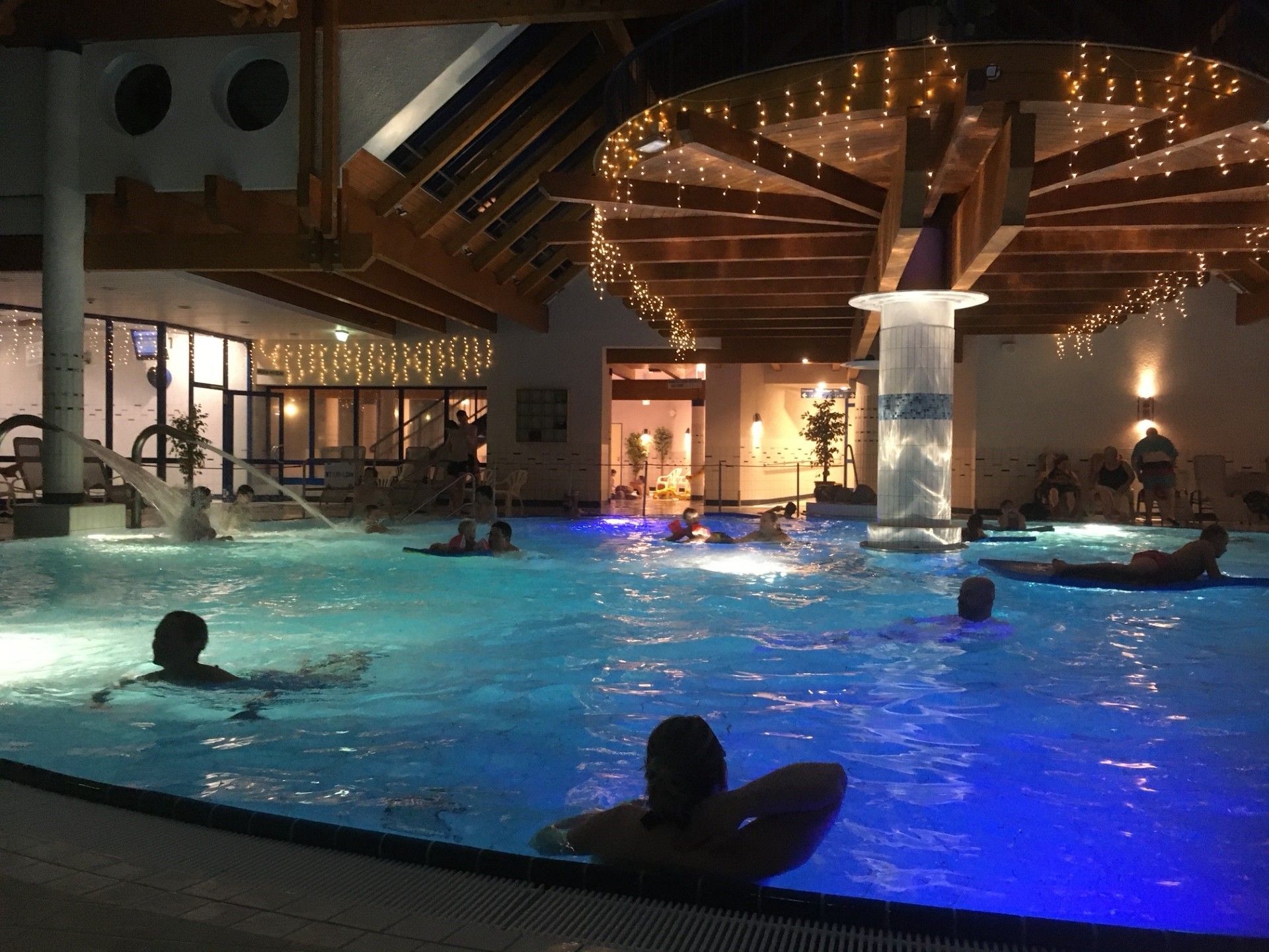 Ein Hallenbad mit mehreren Bahnen, einem überdachten Bereich mit Holzbalken und einer Sitzgelegenheit mit Stühlen.