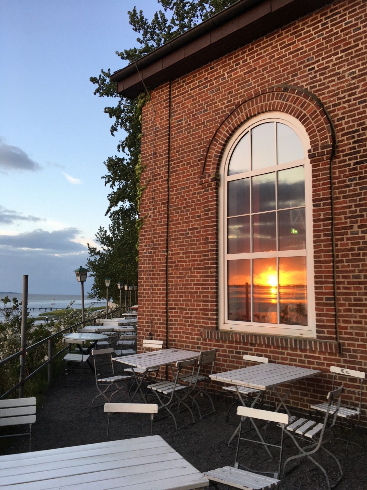 An outdoor seating area with tables and chairs, a brick building with a large arched window, and a view of the sunset over the water.