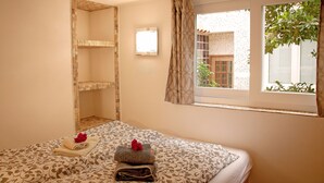 Une chambre d’hôtel avec un lit, une petite fenêtre avec des rideaux, une lampe fixée au mur et une vue sur un bâtiment à l’extérieur.