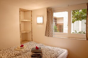 Hotellihuone, jossa on sänky, pieni ikkuna verhoilla, seinään kiinnitetty valaisin ja näkymä ulkona olevaan rakennukseen.