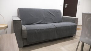 En grå sofa, et rundt bord og en stol i et rum med flisegulv og en dør i baggrunden.