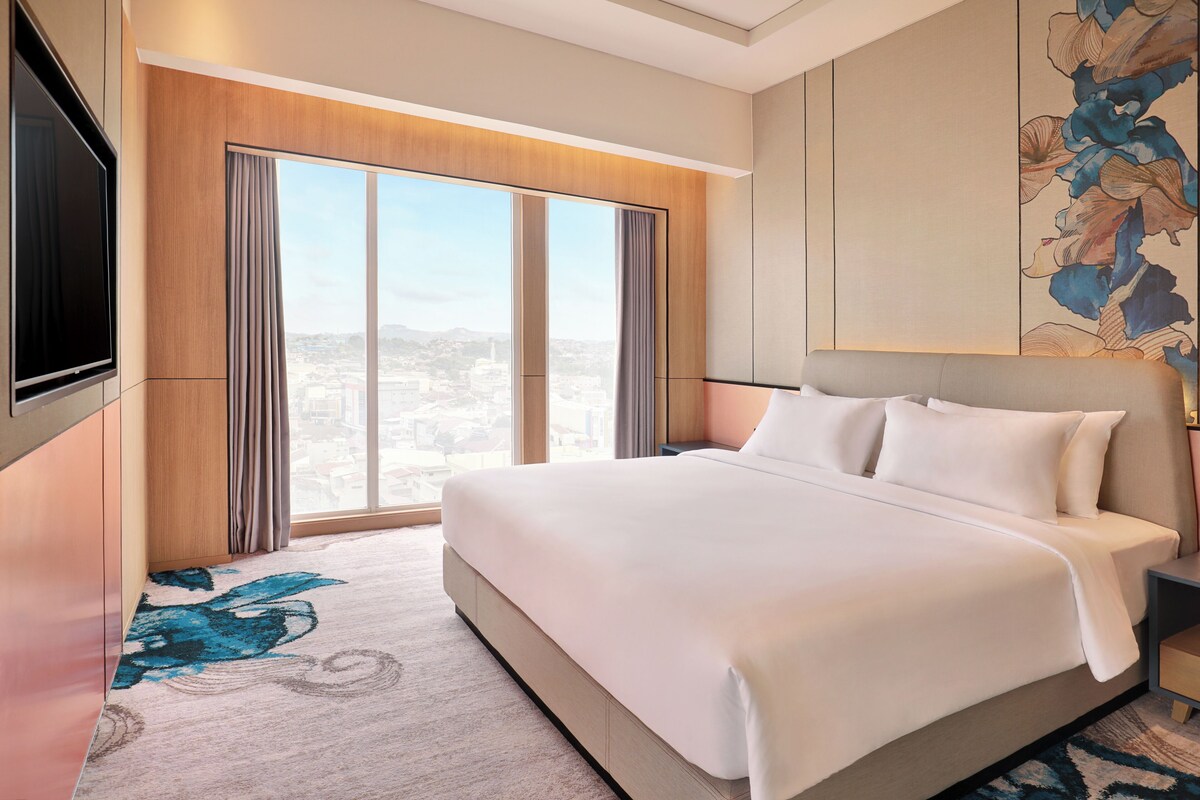 Habitación de hotel con una cama grande, un televisor de pantalla plana y vistas a la ciudad a través de ventanales de gran altura.