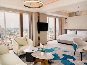 A hotel room with a bed, two armchairs, a small table with a vase, a TV mounted on the wall, and large windows offering a city view. - Mercure Samarinda (Samarinda)