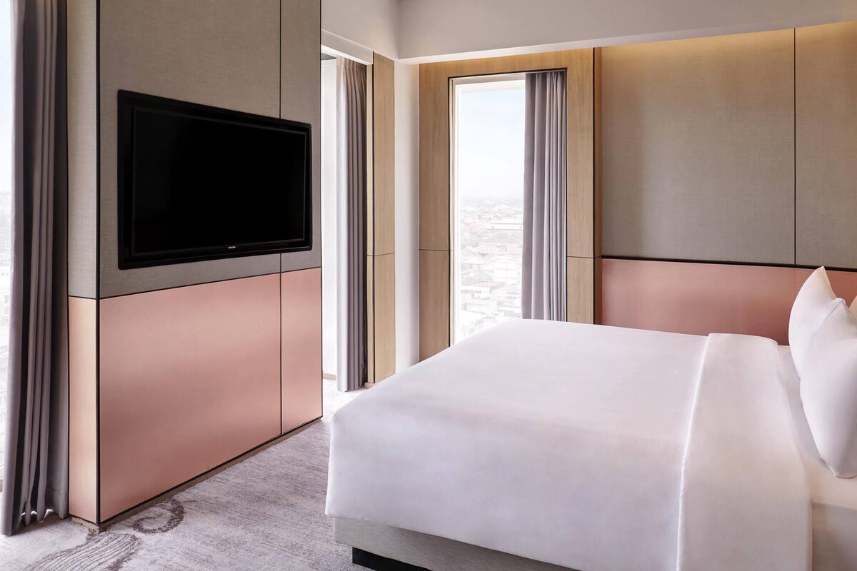 A modern hotel room with a large bed, a flat-screen TV mounted on the wall, and large windows with curtains.
