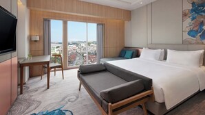 Quarto de hotel com uma cama grande, um sofá, uma mesa de jantar com cadeiras, uma televisão e vista para a cidade pela janela.