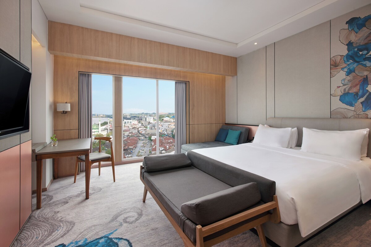 Habitación de hotel con una cama grande, un sofá, una mesa de comedor con sillas, un televisor y vistas a la ciudad desde la ventana.