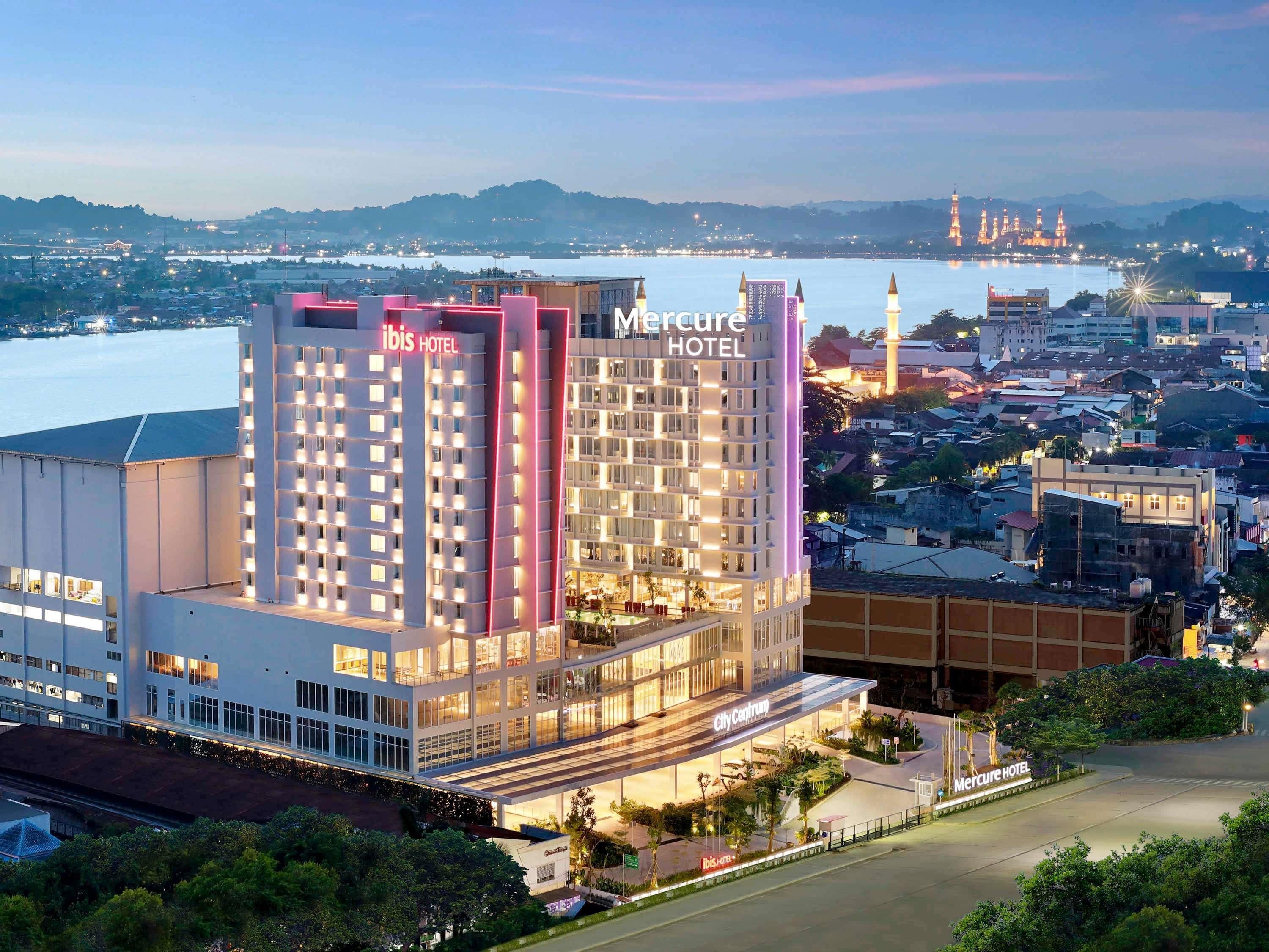 A modern hotel with a prominent 'Mercure Hotel' sign, a spacious lobby, and a view of the cityscape and water in the background.