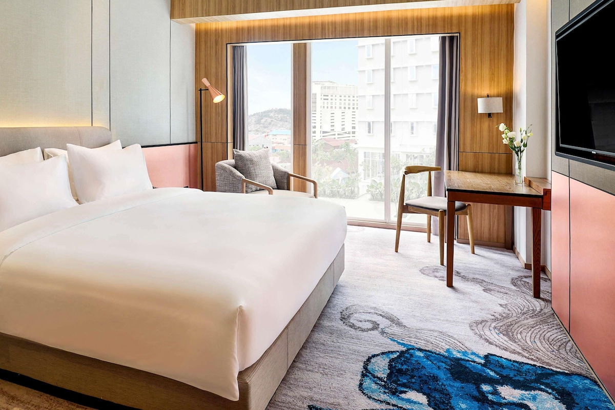 Habitación de hotel con una cama grande, un escritorio con silla, un televisor y vistas a edificios a través del ventanal.