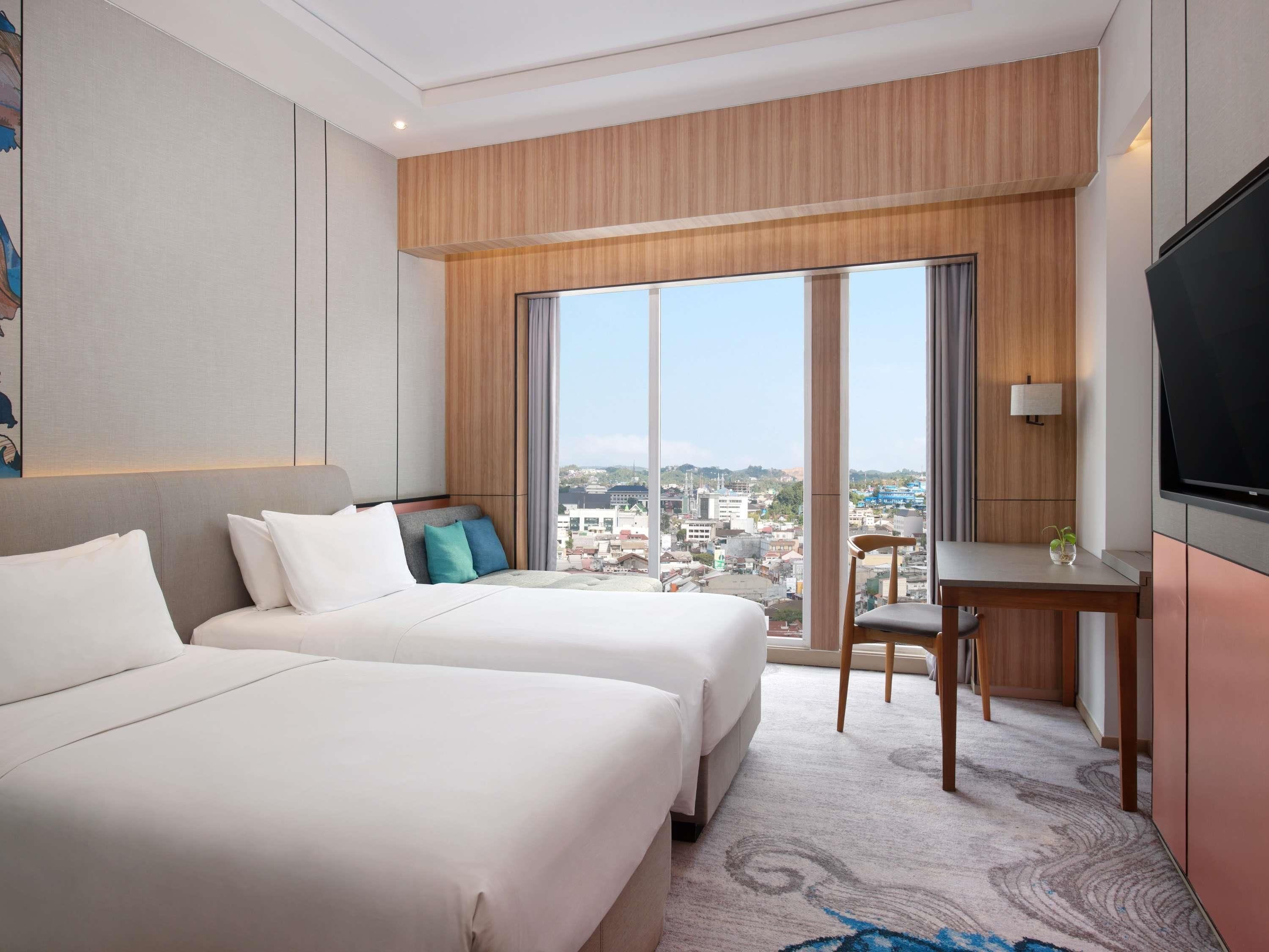 A hotel room with two beds, a desk, a chair, a TV, and a large window with a city view.
