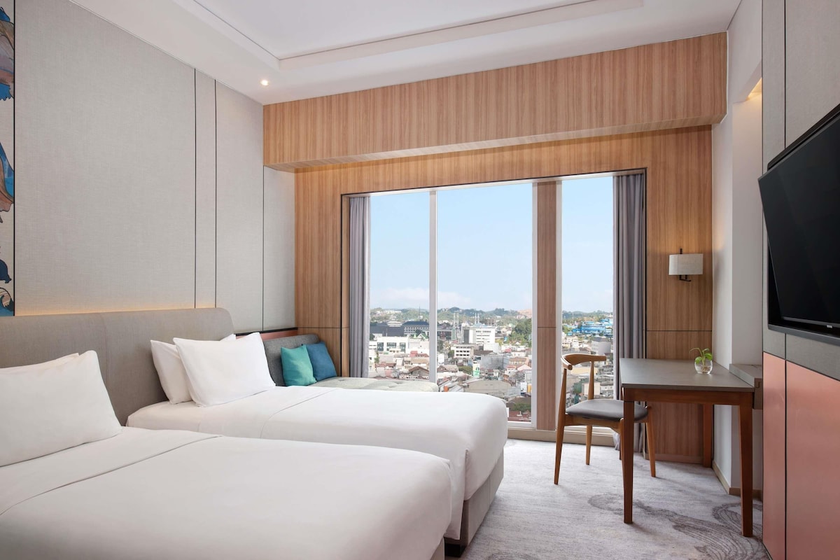 Habitación de hotel con dos camas, un escritorio, una silla, un televisor y un ventanal con vistas a la ciudad.