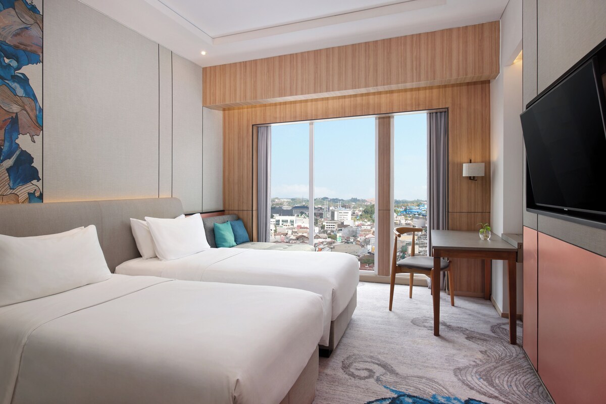 Habitación de hotel con dos camas, un escritorio, una silla, un televisor y un ventanal con vistas a la ciudad.