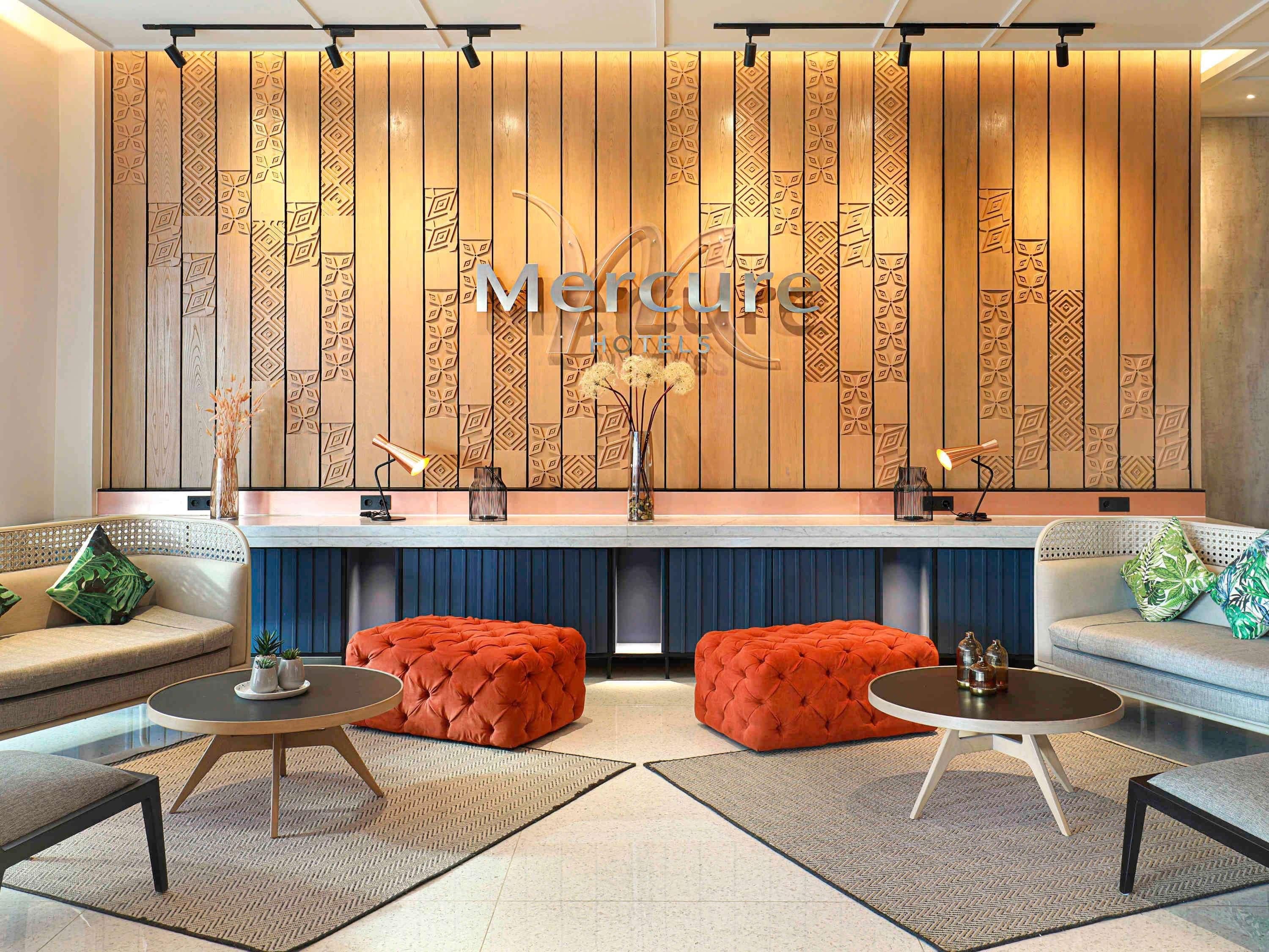 A modern living room with a wooden panel wall, two orange ottomans, a round coffee table, and a sofa with patterned cushions.
