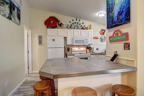 A kitchen with white cabinets, a microwave, a refrigerator, and a large island with stools. There are decorative items on the walls and a sign that says "BEACH HOUSE".