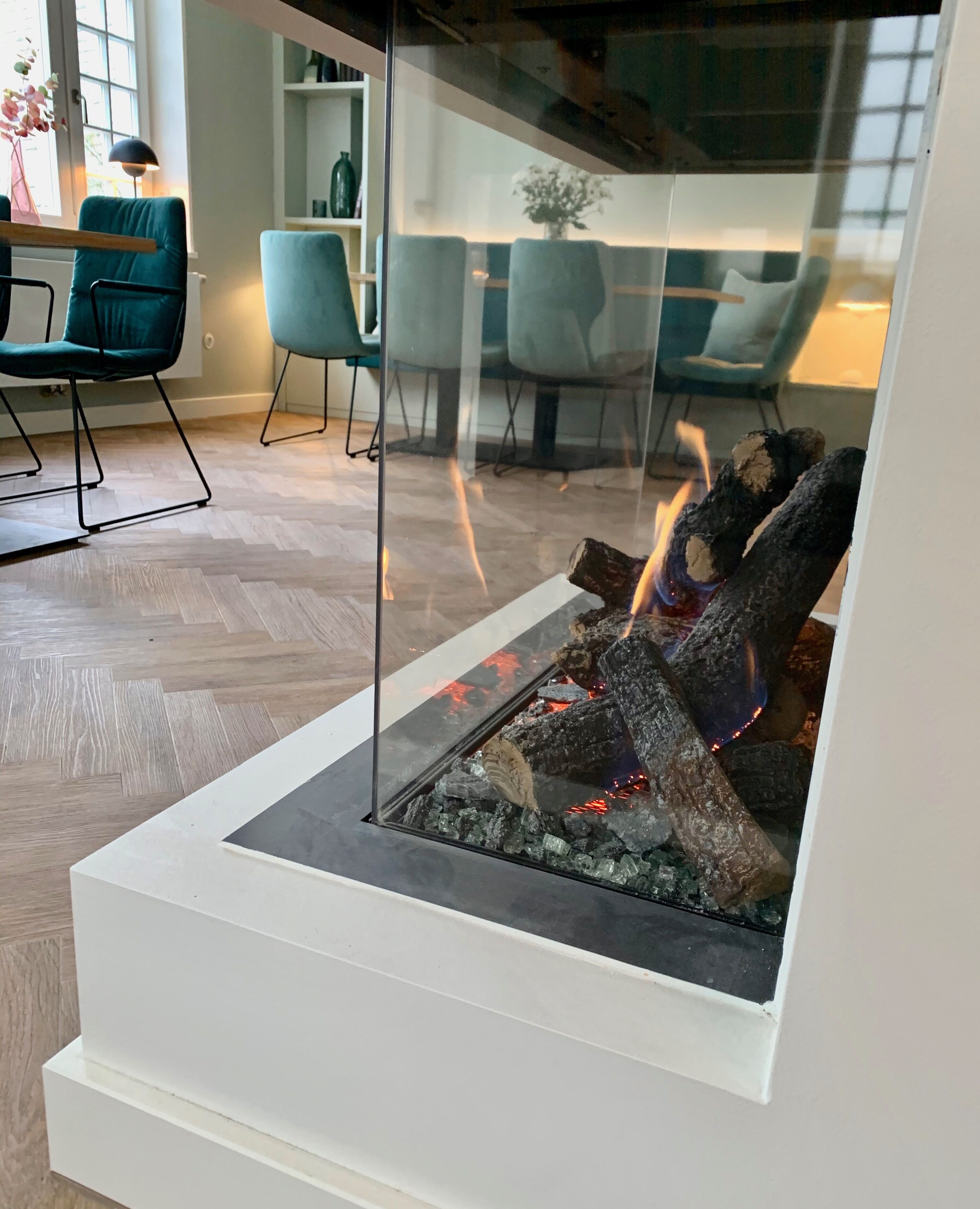 Un salon moderne doté d’une cheminée à façade en verre, de planchers en bois et de fauteuils de couleur turquoise.