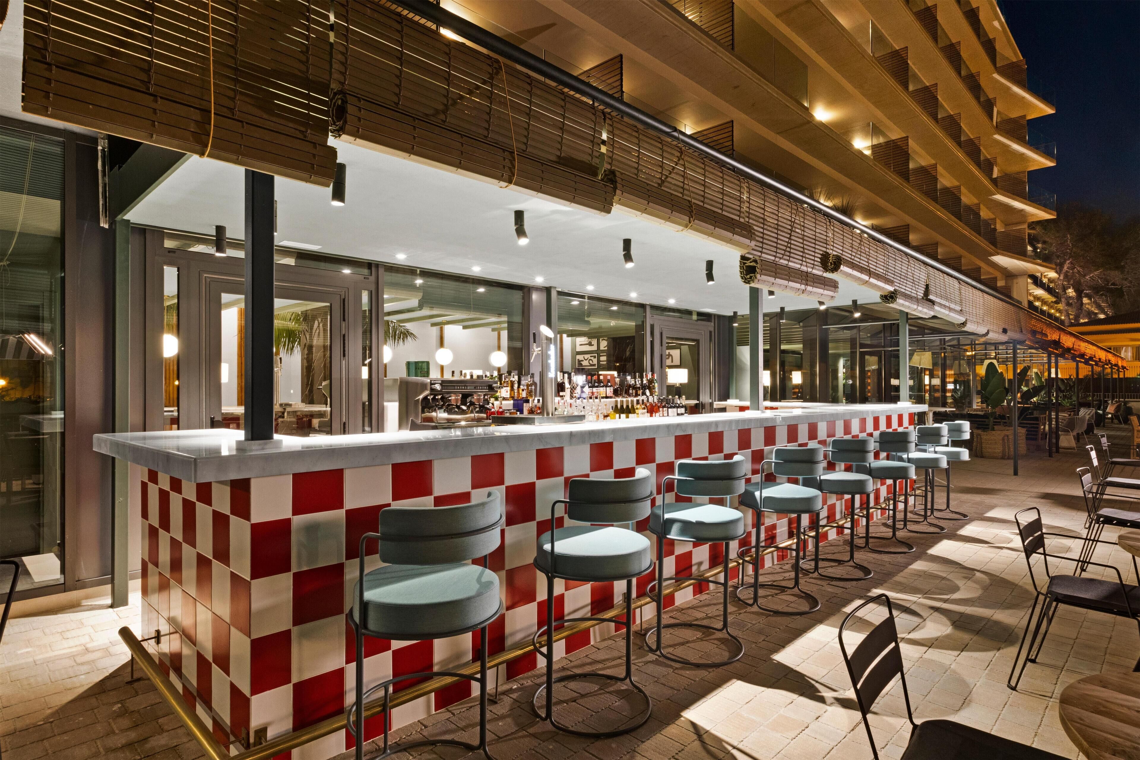 Zona de bar con suelo a cuadros rojo y blanco, varios taburetes y una barra con cerramiento de vidrio.