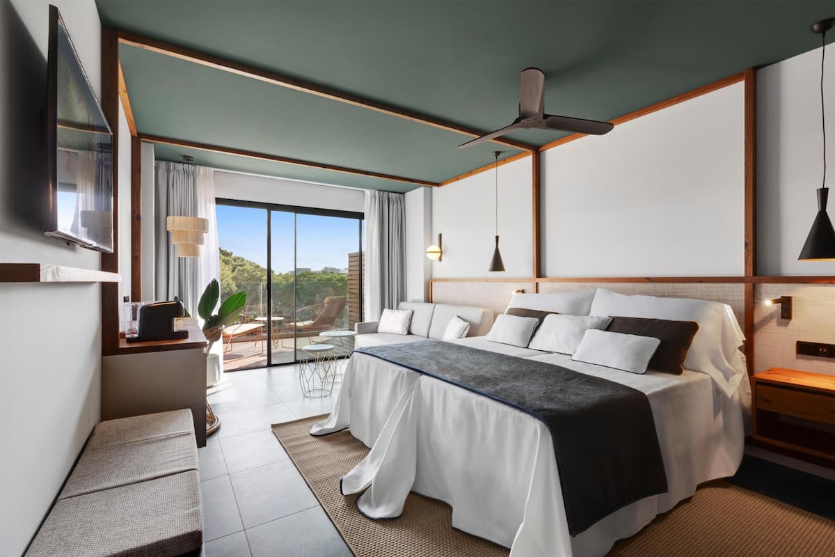 Habitación de hotel moderna con una cama grande, un escritorio, una silla, un televisor y un balcón con vistas a la ciudad.
