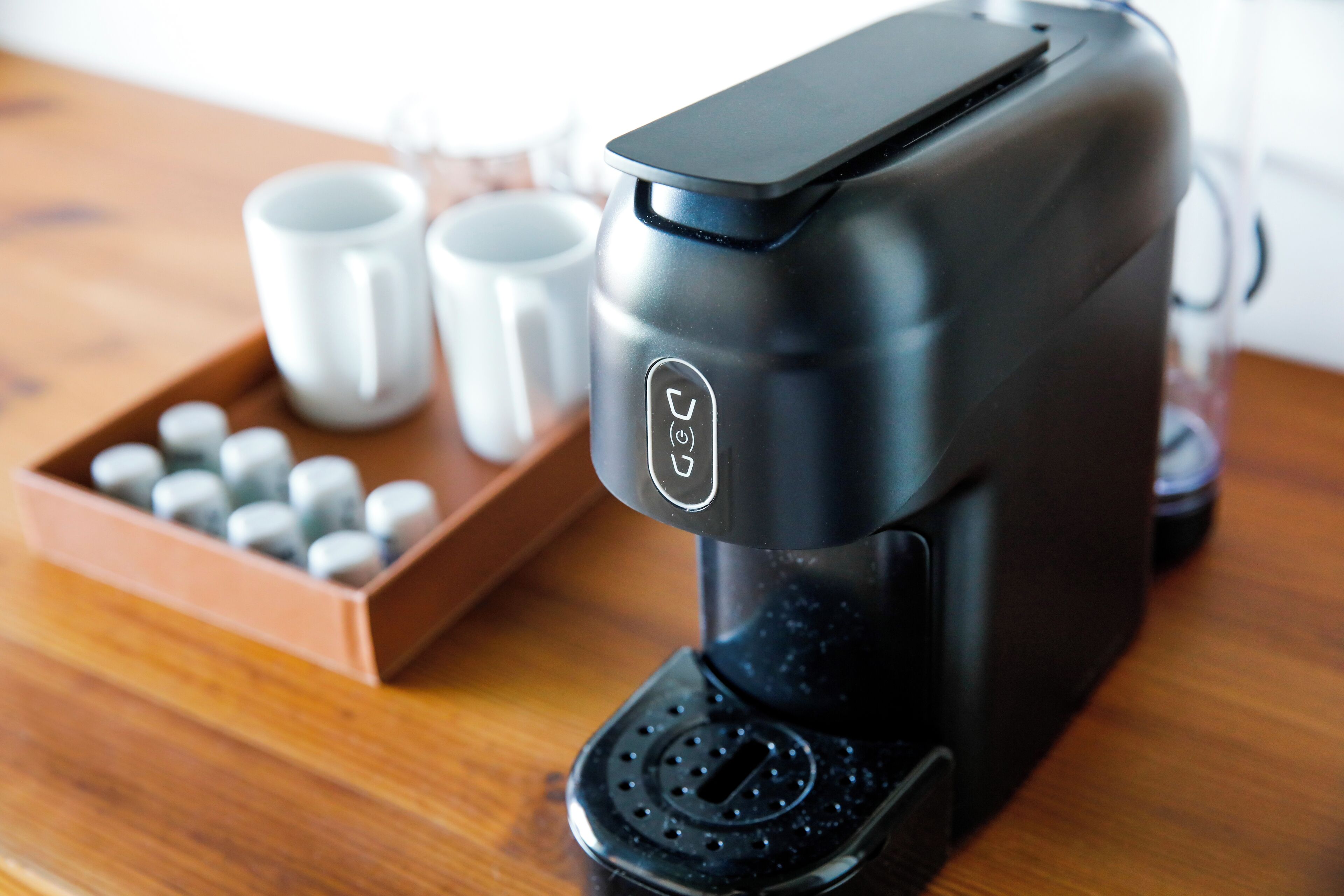 Una cafetera con panel de control, una bandeja con cápsulas y dos tazas blancas sobre una mesa de madera.