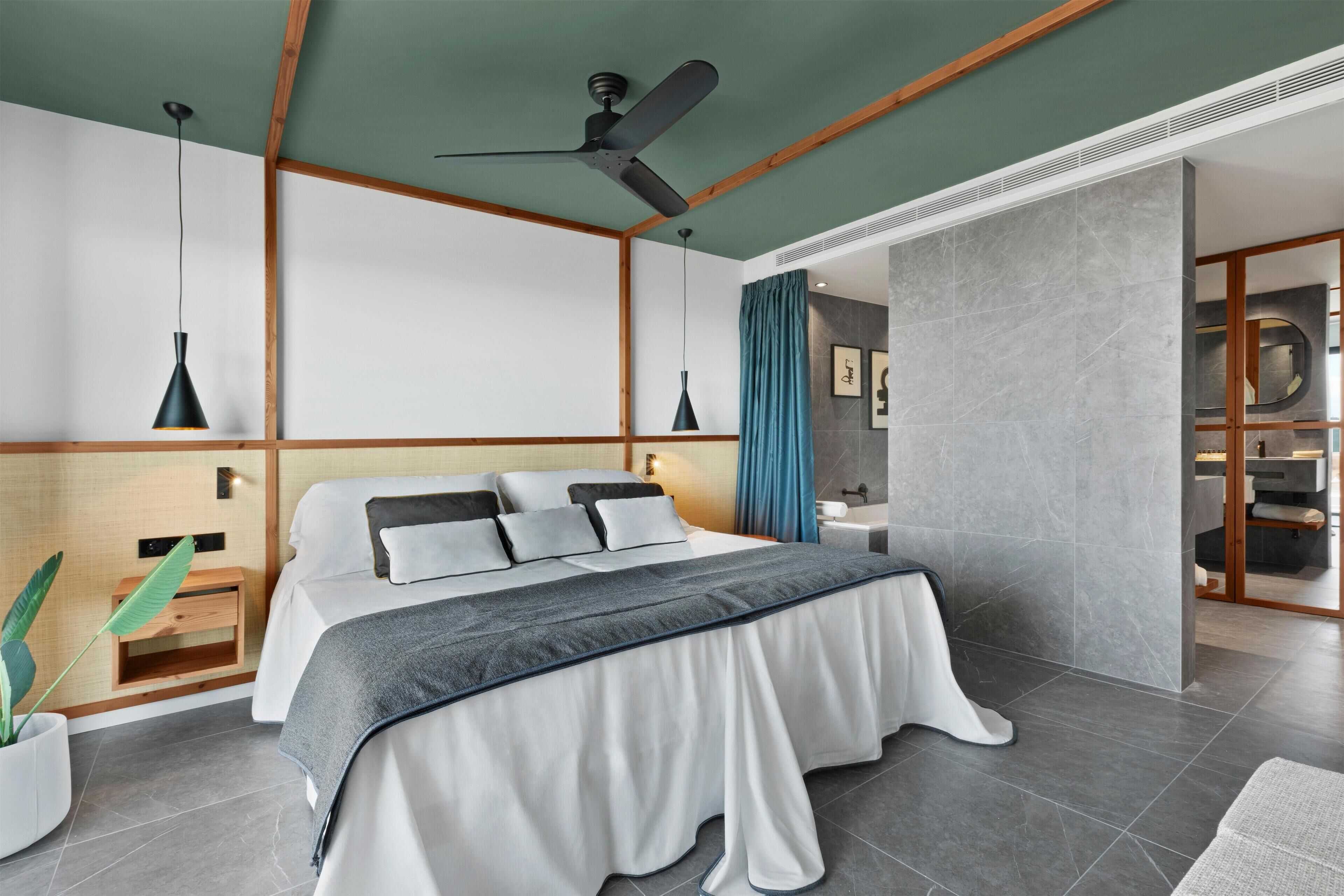 Quarto moderno com cama, mesas de cabeceira, ventoinha no teto e banheiro visível por uma porta aberta.