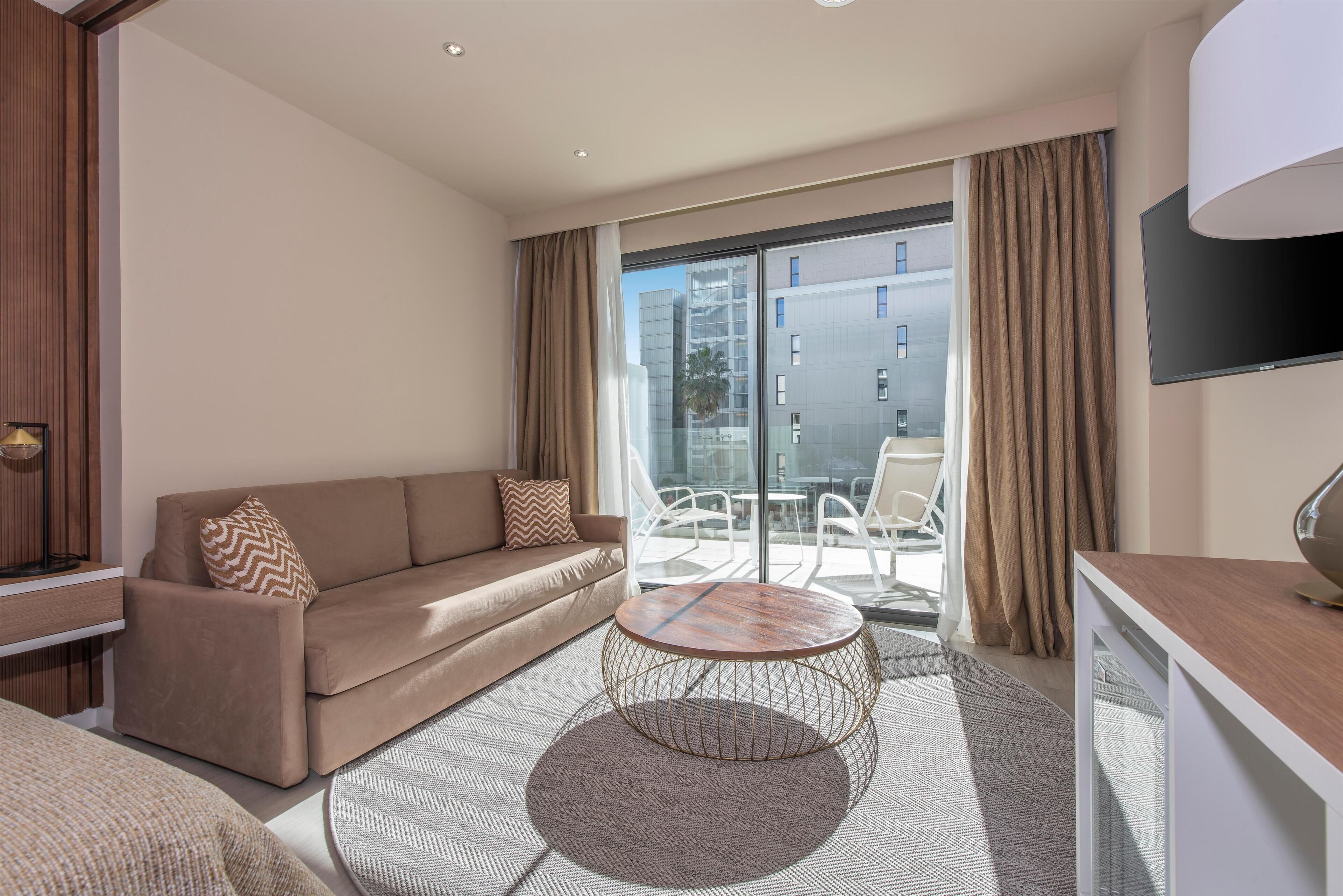 Una habitación de hotel moderna con sofá, una mesita redonda, televisión de pantalla plana y balcón con vistas a la ciudad.
