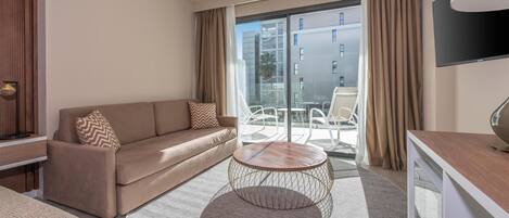 A modern hotel room with a sofa, a round coffee table, a flat-screen TV, and a balcony with a view of the city.