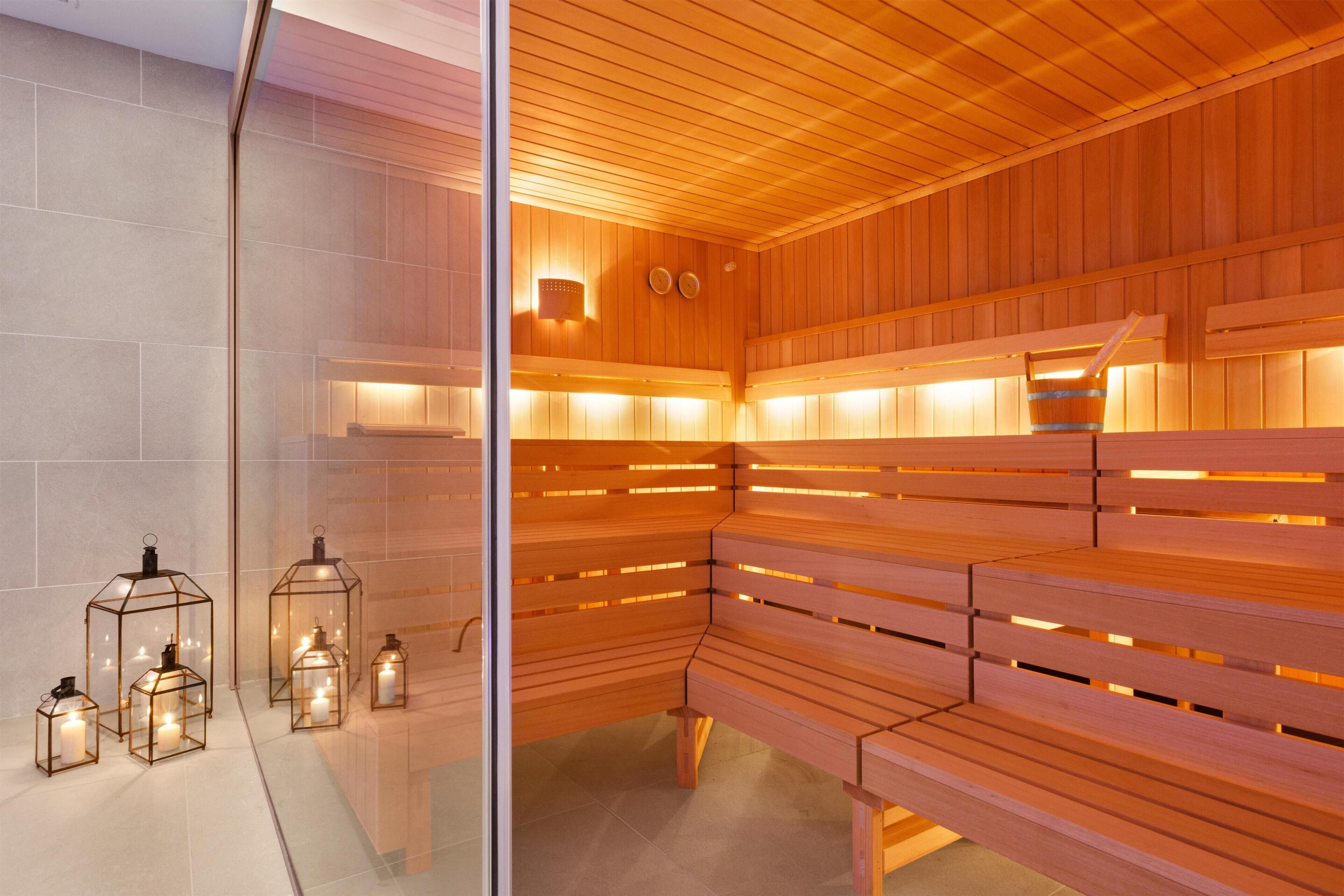 A sauna with wooden benches and glass doors, featuring wall-mounted heaters and candle-like lights.