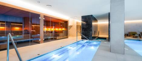 A modern indoor swimming pool with clear blue water, glass walls, and wooden seating areas.