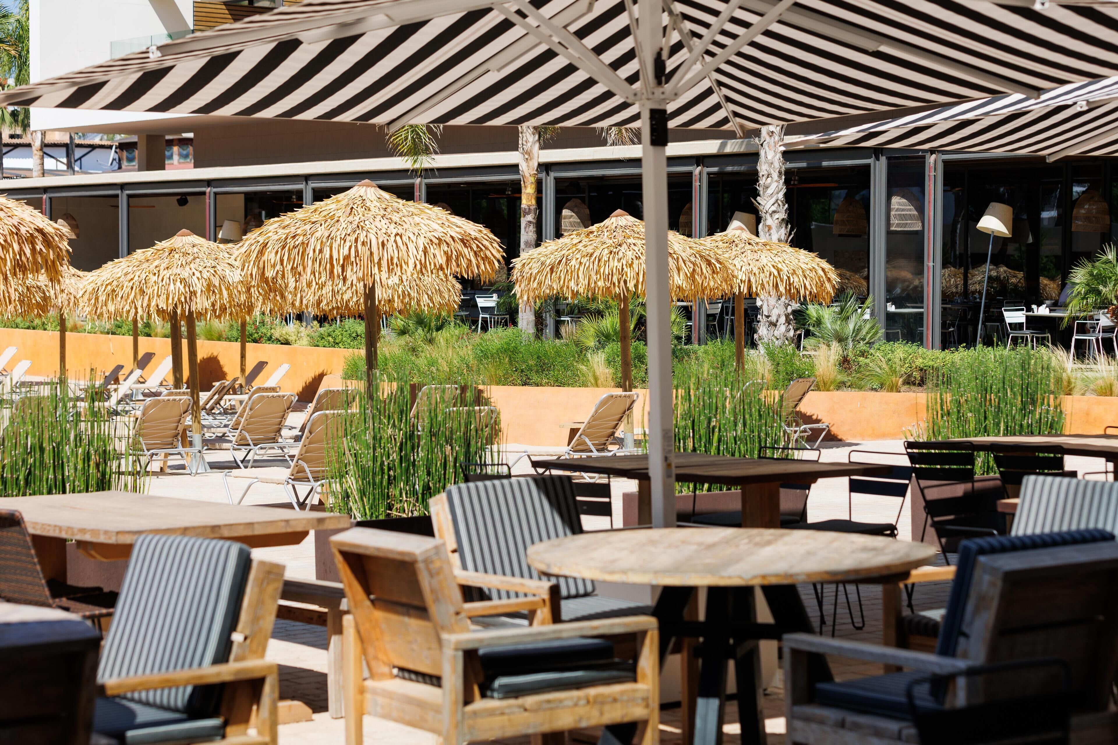 An outdoor seating area with round tables and chairs, thatched umbrellas, and a modern building in the background.