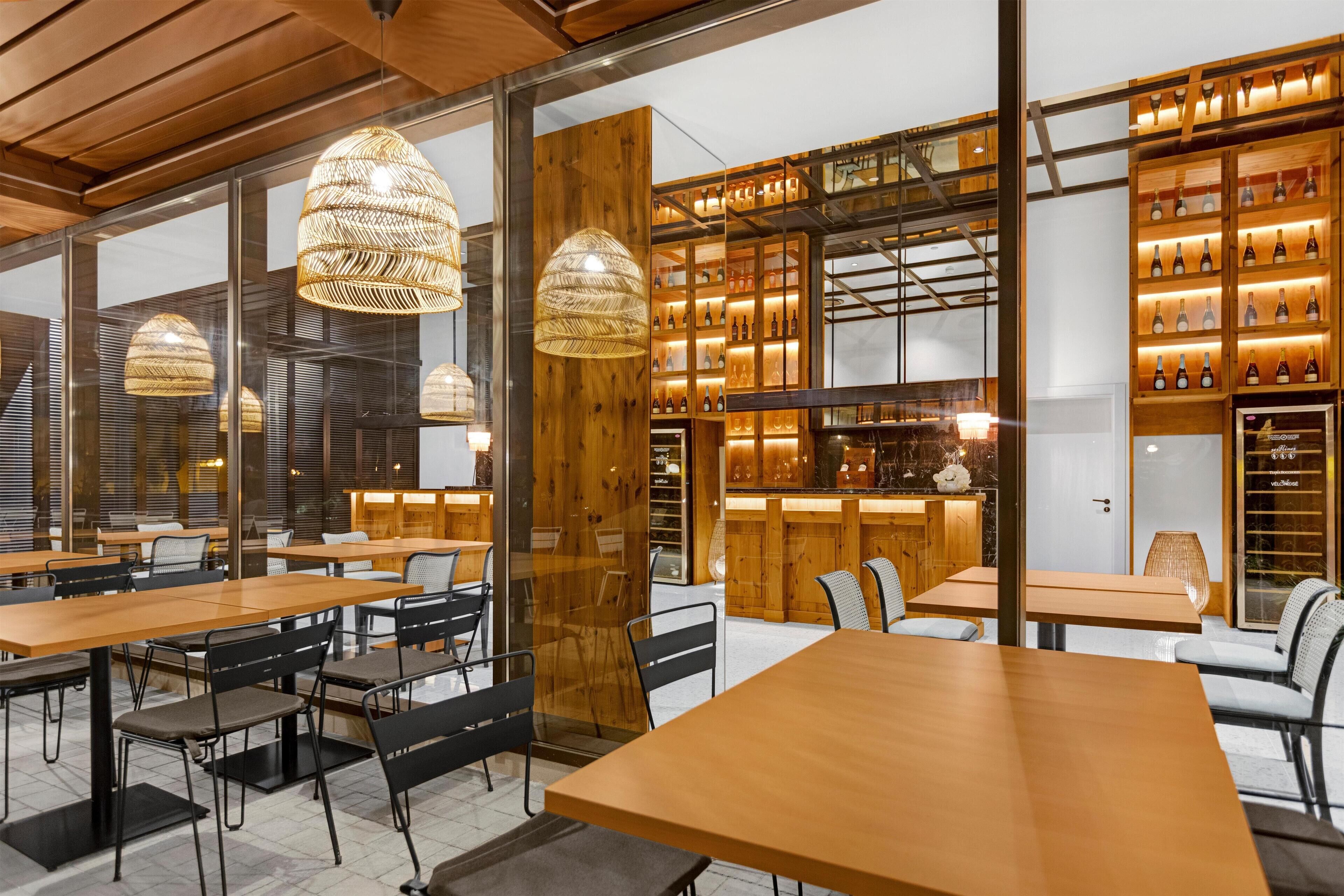 A modern restaurant with wooden tables and chairs, hanging rattan lamps, and a wine display.