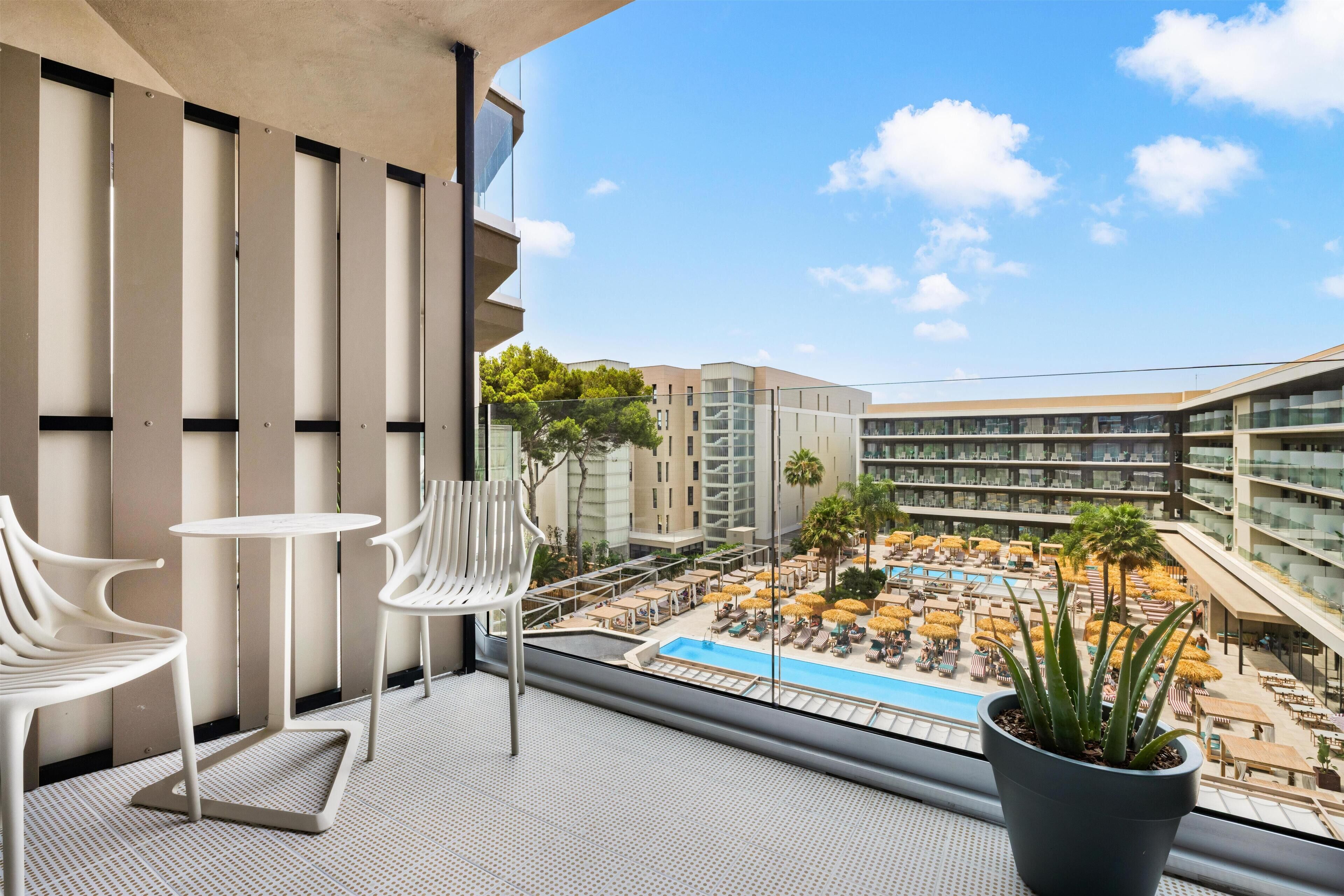 A balcony with white chairs and a potted plant, overlooking a pool area with umbrellas and lounge chairs.