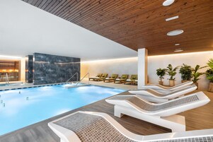 Un espace piscine intérieur moderne, doté de chaises longues, d’un mur en pierre et d’un plafond en bois.