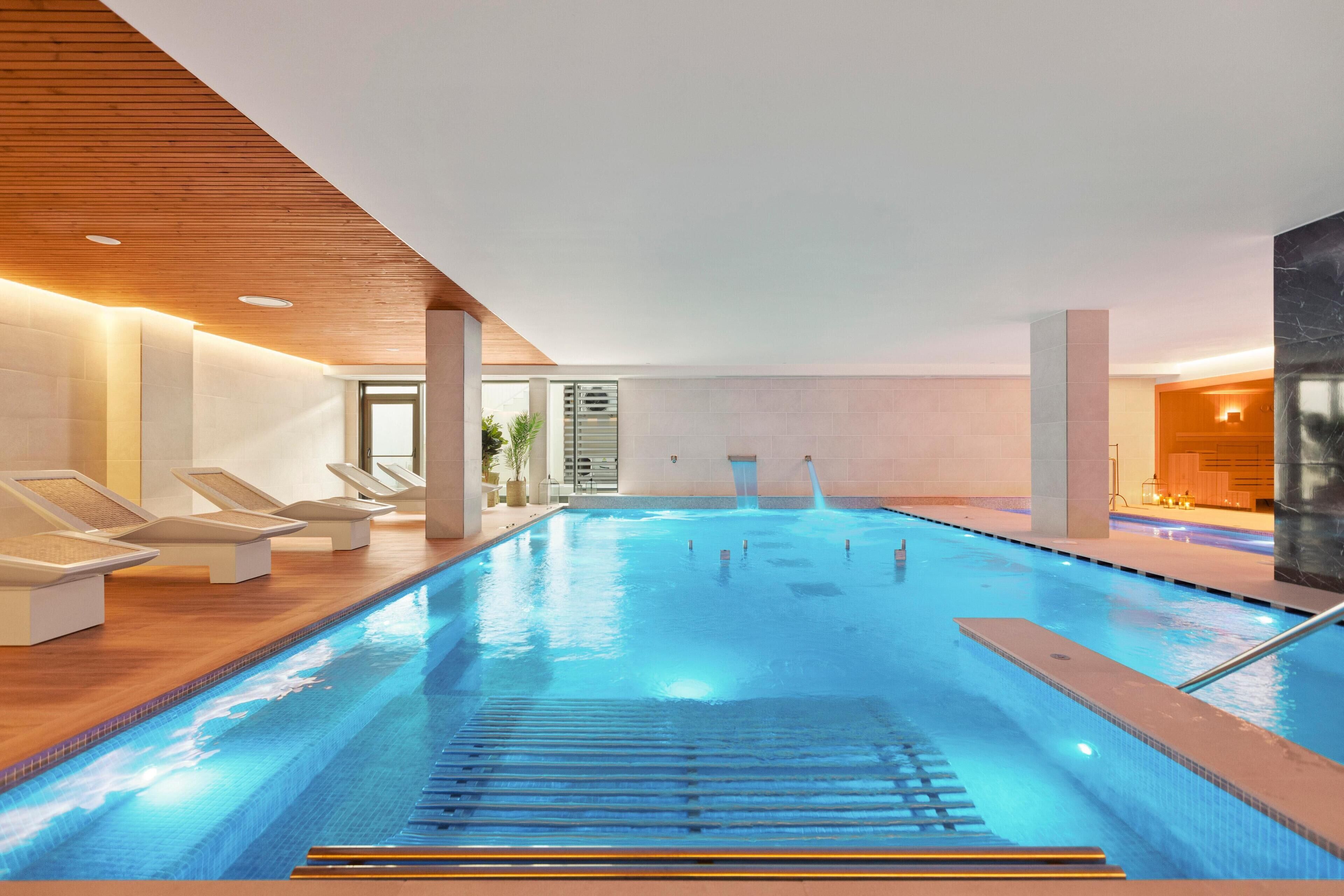 An indoor swimming pool with clear water, sun loungers, and a wooden ceiling.