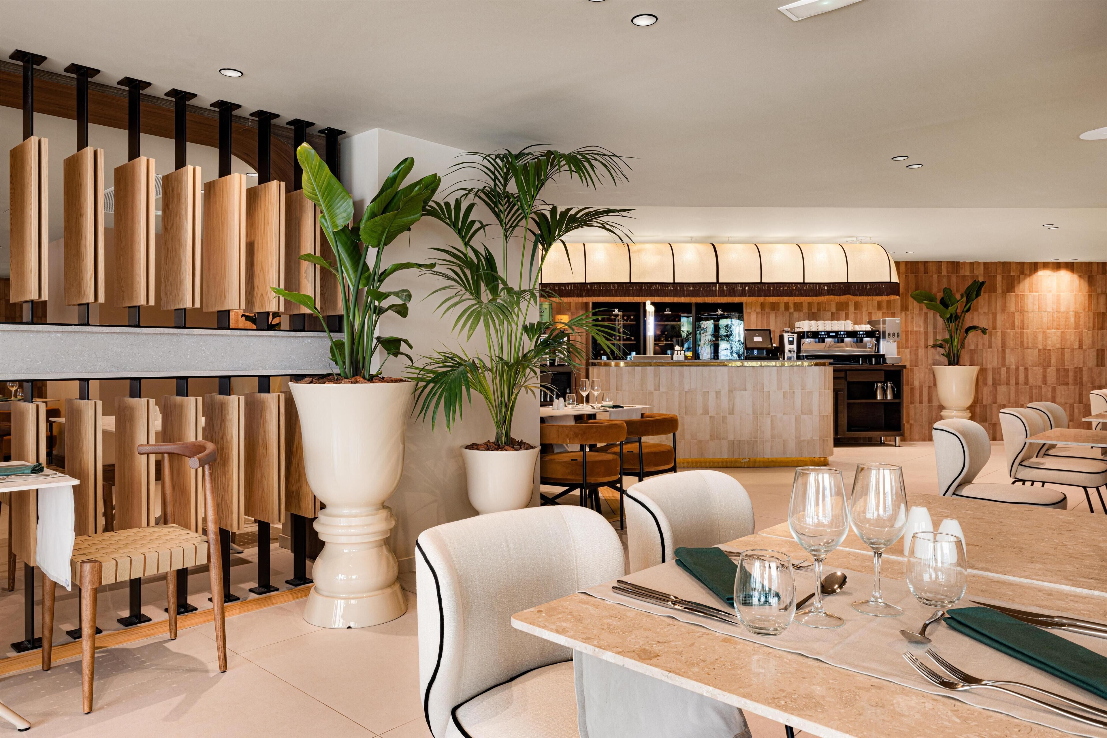 A modern restaurant with a bar area, wooden chairs, potted plants, and a marble-topped dining table.