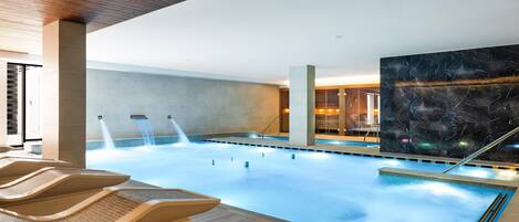 Una piscina interior moderna con superficie de agua cristalina, techo de madera y una amplia pared de mármol.