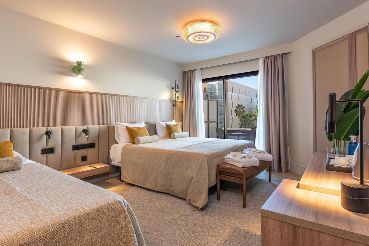 Habitación de hotel con dos camas, una mesita de noche, un escritorio con lámpara, un ventanal con cortinas y vistas al exterior.