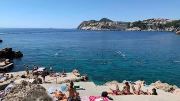 A coastal beach scene with people sunbathing and swimming, rocky terrain, and buildings in the background.