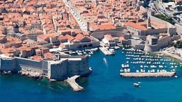 An aerial view of a coastal city with a prominent fortress, a marina with numerous boats, and densely packed buildings with red-tiled roofs.