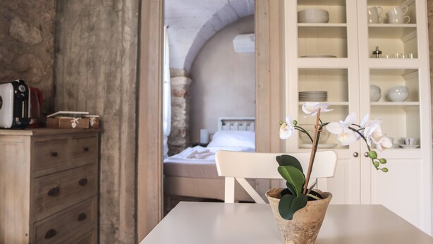 Una habitación con un aparador de madera, una mesa blanca con una orquídea en maceta y un mueble blanco con vajilla y tazas.