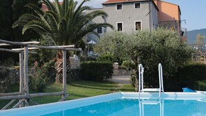 Una piscina con escalera, rodeada de vegetación y un edificio con techo rojo al fondo.